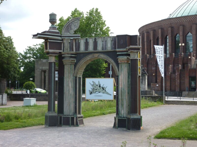 Blick in DIE GROSSE 2022. Außenskulptur: Bogen von Clemens Botho Goldbach. Weiße Stellwände von Ivo Weber mit 