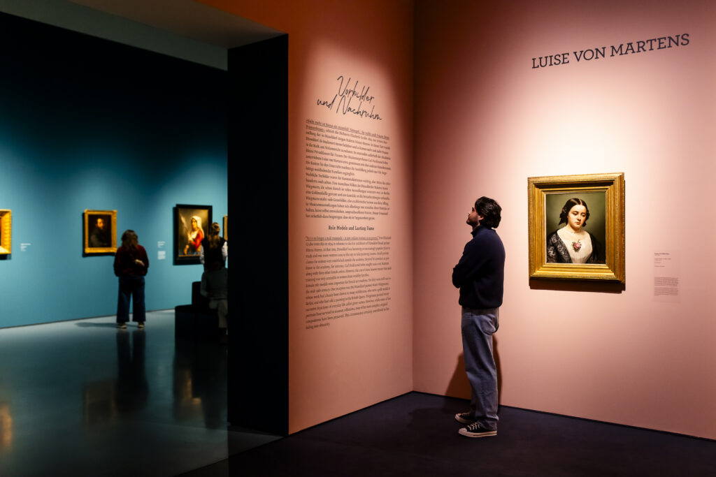 Ein Mann steht in einer Kunstgalerie und liest einen Text an der Wand neben einem Porträt mit der Aufschrift Luise von Martens. Andere Besucher betrachten die Kunst in der Nähe.