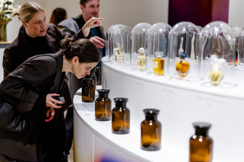 Zwei Frauen untersuchen und riechen Düfte aus braunen Glasflaschen in einer Parfümausstellung, mit Glaskuppeln im Hintergrund.