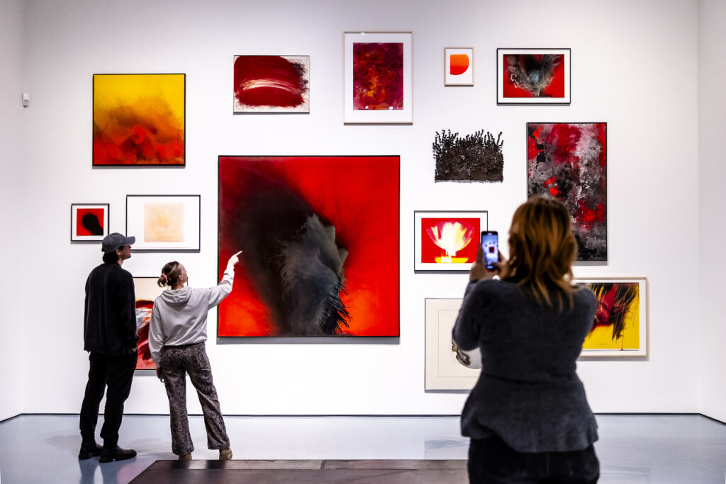 Drei Personen betrachten und fotografieren bunte abstrakte Kunstwerke, die auf einer weißen Galeriewand ausgestellt sind.