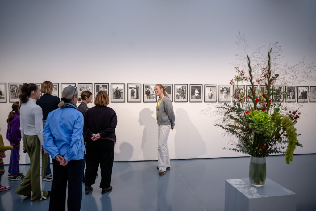 Eine Gruppe von Menschen betrachtet Schwarz-Weiß-Fotografien an einer Galeriewand; im Vordergrund steht ein großes Blumenarrangement.