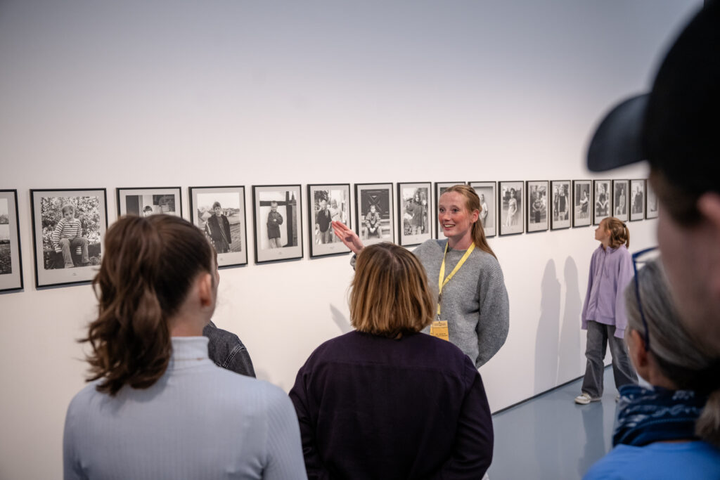 Eine Frau führt eine Gruppe durch eine Kunstgalerie und bespricht eine Reihe von Schwarz-Weiß-Fotografien, die an der Wand hängen.