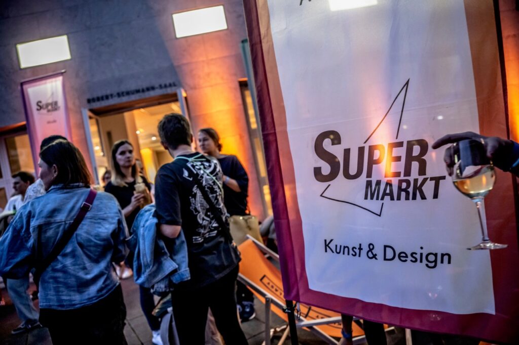 Menschen versammeln sich bei einer Kunst- und Designveranstaltung im Freien mit einem Super Markt Kunst & Design-Banner und jemandem, der ein Glas Wein hält.
