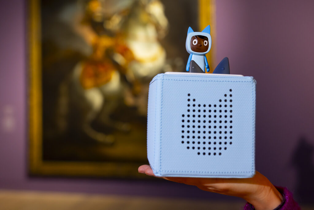 Eine Hand hält eine blaue Audiobox mit einem Katzengesicht und einer kleinen Figur mit Katzenohren auf der Oberseite, mit Kunst im verschwommenen Hintergrund.
