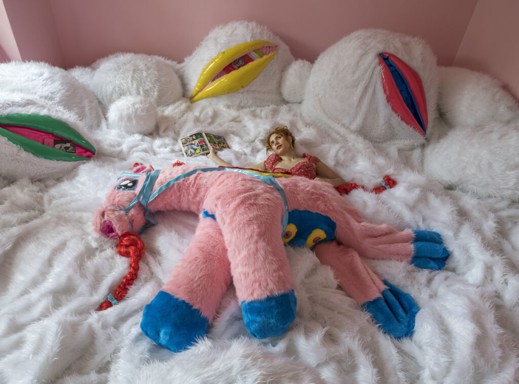 Eine Person in Rosa liegt auf einem großen, flauschigen Bett mit Plüschlippen und einem riesigen rosa Plüschpferd und liest eine Zeitschrift.