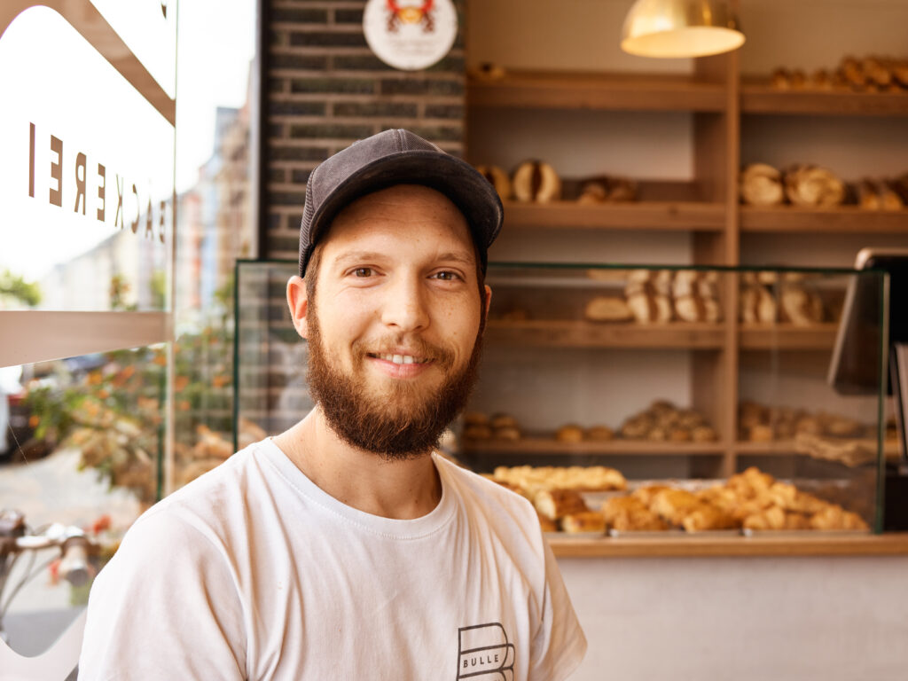 Ein lächelnder Mann mit Bart und Mütze steht in einer Bäckerei, mit Regalen voller Brot und Gebäck im Hintergrund.