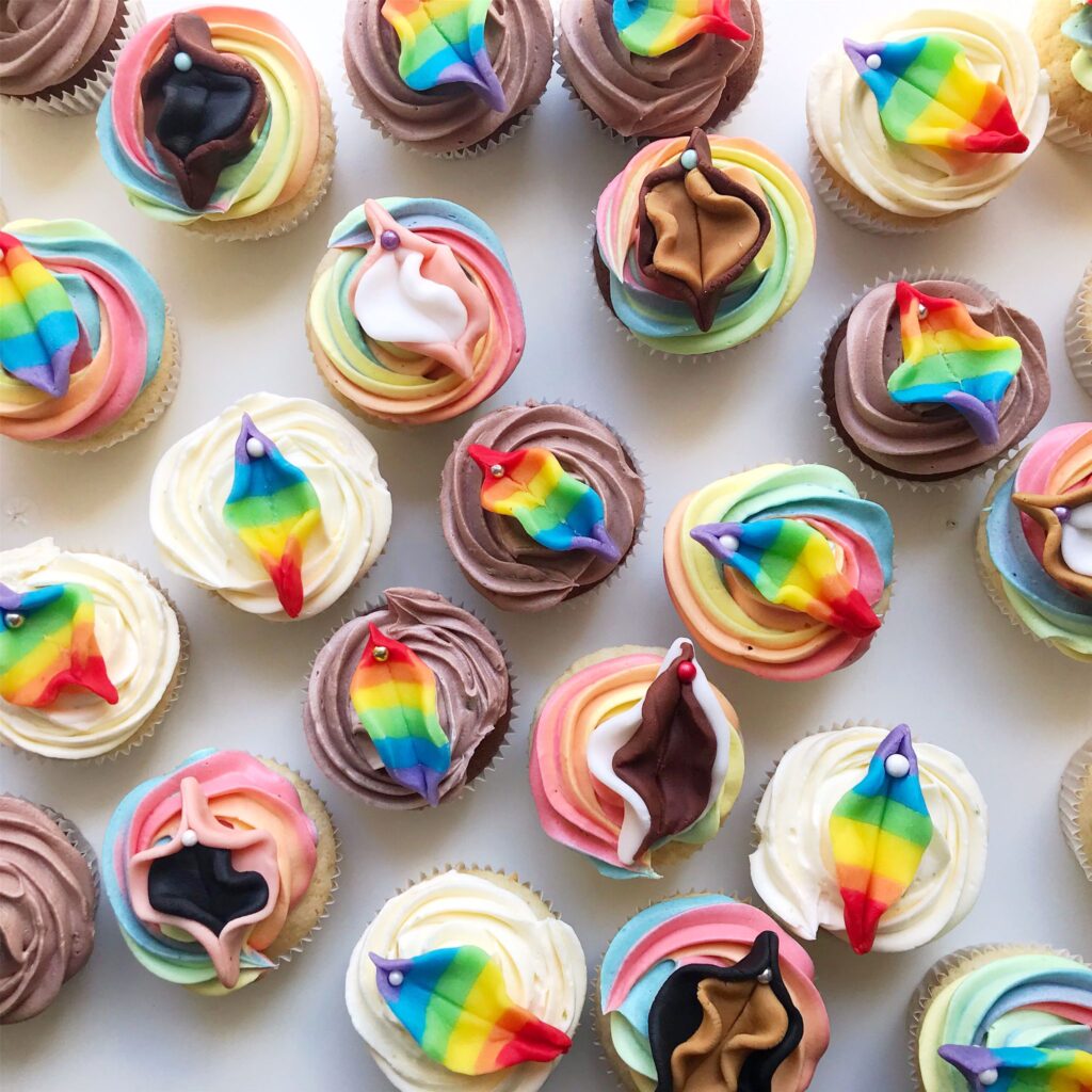 Verschiedene Cupcakes mit regenbogen- und vulvenförmigen Verzierungen auf pastellfarbenen und schokoladenfarbenen Tortenböden, angeordnet auf einer weißen Fläche.