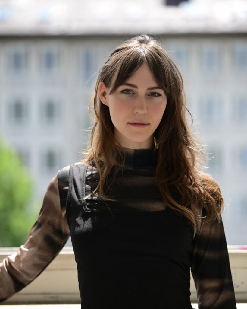 Eine Frau mit langen braunen Haaren und einem durchsichtigen schwarzen Oberteil steht an einem Fenster mit einem verschwommenen Gebäude im Hintergrund.