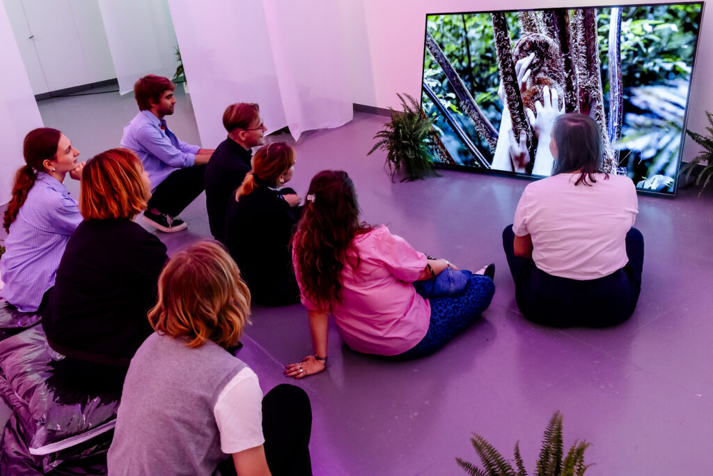 Acht Personen sitzen auf dem Boden vor einem großen Bildschirm, auf dem Hände zu sehen sind, die Äste in einem Wald berühren.