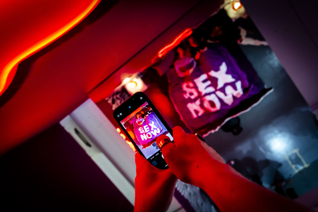 Eine Person macht ein Foto von einem T-Shirt mit der Aufschrift $EX NOW in einem rot und lila beleuchteten Raum.