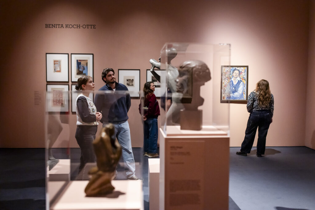 Man betrachtet Skulpturen und gerahmte Kunstwerke in einer Museumsgalerie mit rosa Wänden und BENITA KOCH-OTTE an der Wand.