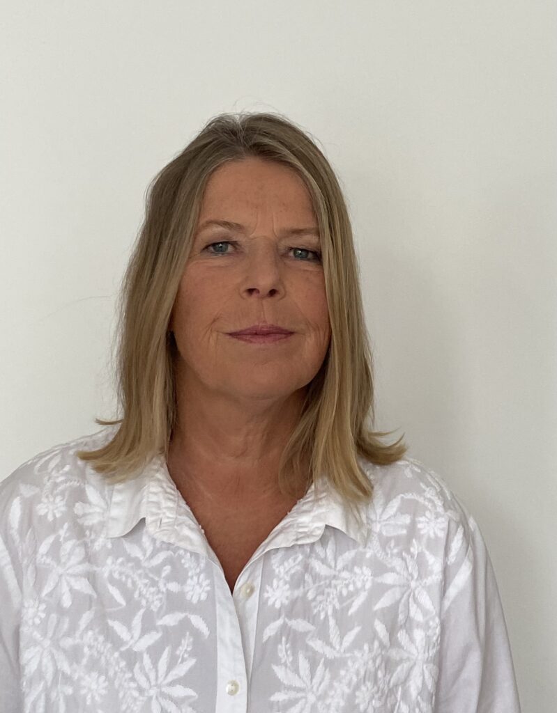 Eine Frau mit schulterlangem blondem Haar trägt eine weiße, blumengemusterte Bluse und steht vor einem schlichten weißen Hintergrund.