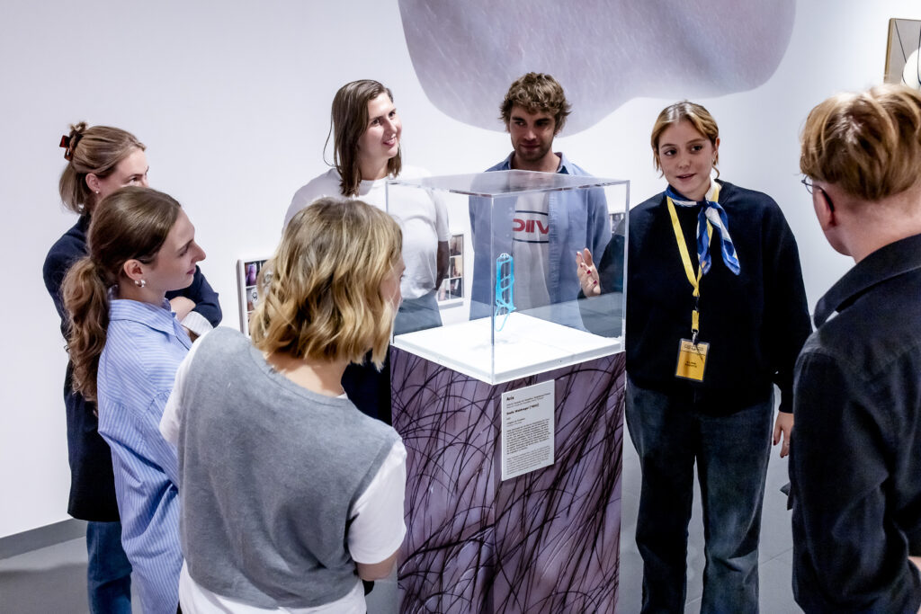 Eine Gruppe von Menschen steht um eine Vitrine in einer Galerie und hört einer Frau mit einem Schlüsselband zu, die die Ausstellung erklärt.