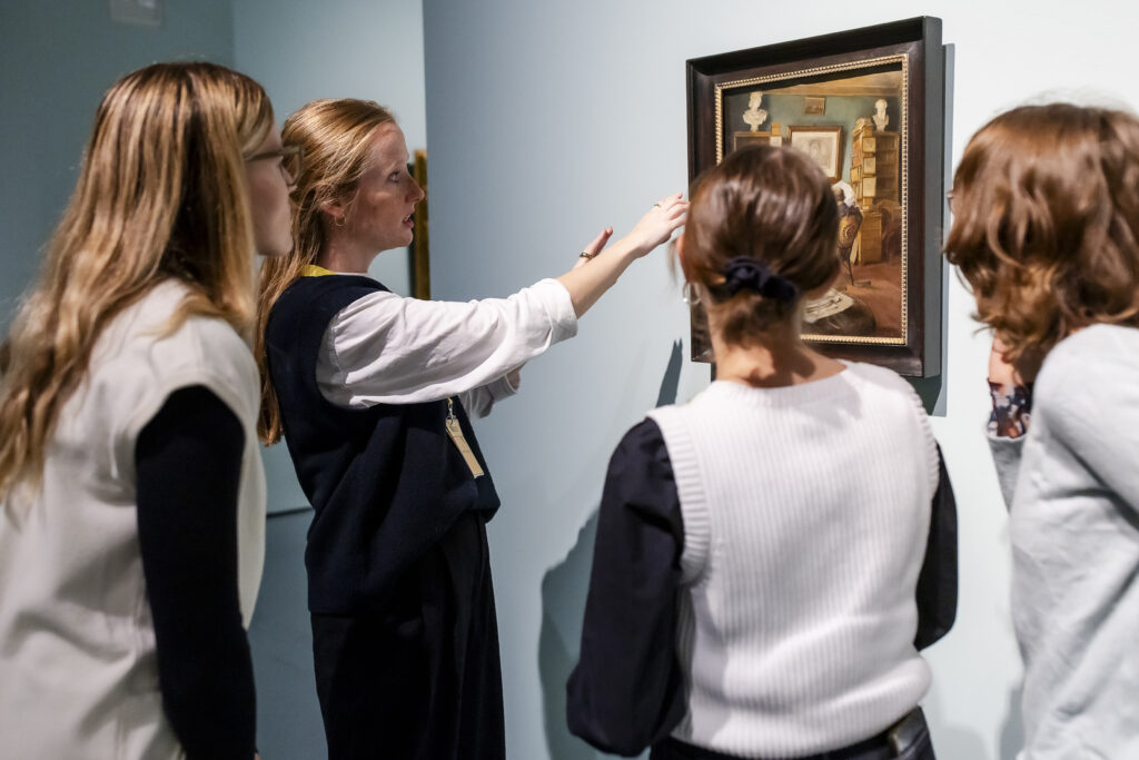 Vier Frauen stehen dicht beieinander und betrachten und besprechen ein gerahmtes Gemälde an einer Galeriewand.