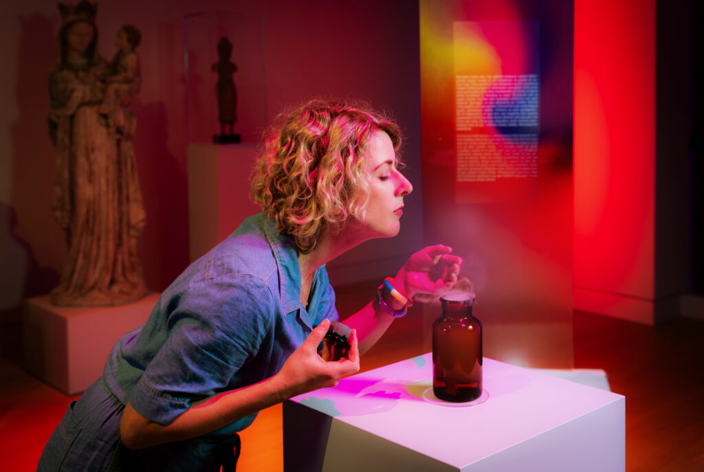 Eine Frau mit lockigem Haar riecht an einer dunklen Glasflasche, die in einem farbenfrohen, modernen Kunstmuseum ausgestellt ist.