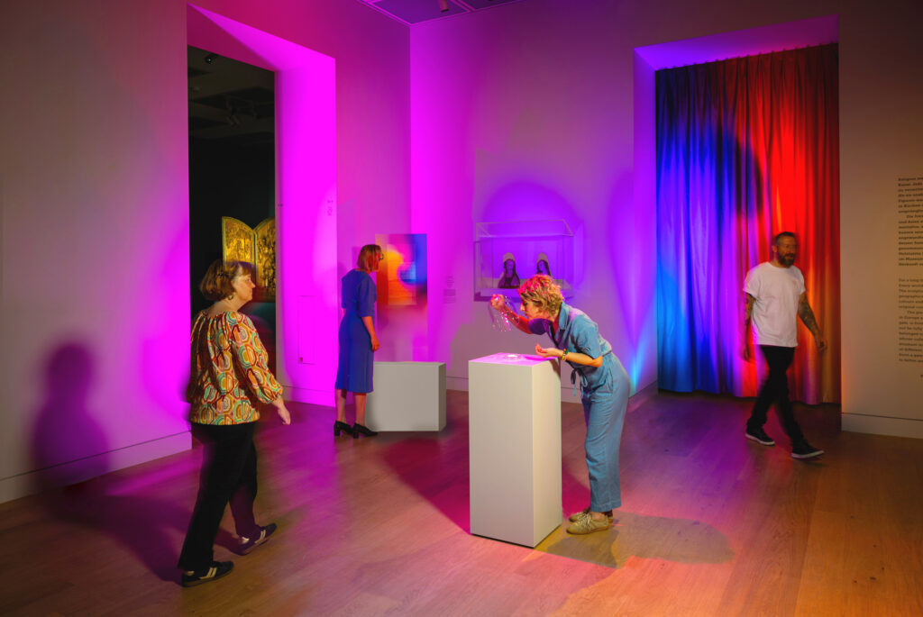 Vier Personen erkunden eine farbenfrohe Kunstausstellung mit dramatischer rosa, lila und blauer Beleuchtung in einem modernen Galerieraum.
