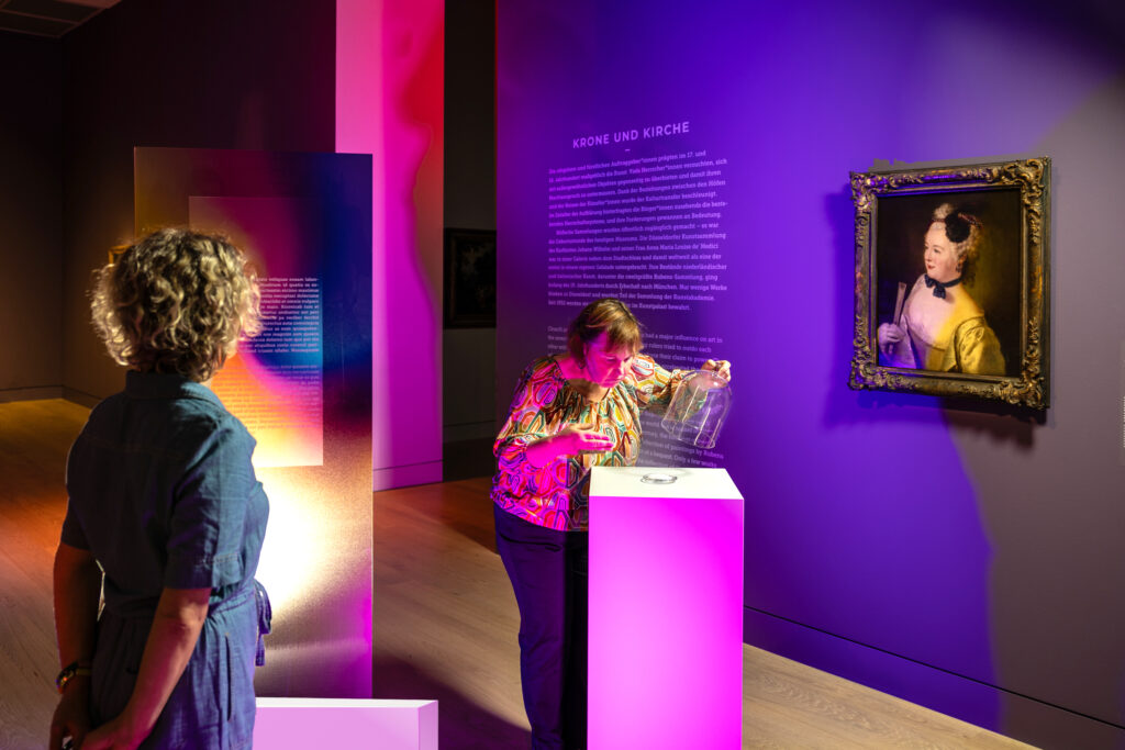 Zwei Frauen betrachten eine Kunstausstellung; eine gießt Wasser auf ein Podest, bunte Lichter erhellen die Szene, und in der Nähe hängt ein gerahmtes Porträt.