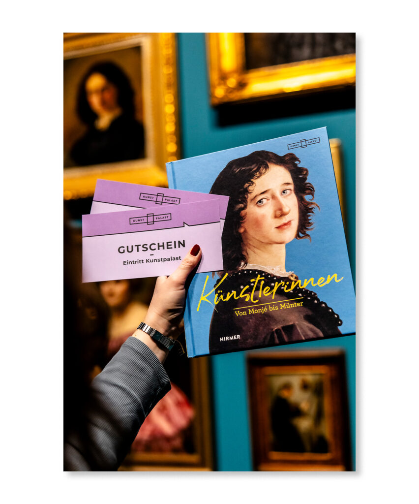 Eine Hand hält Museumsgutscheine und das Buch Künstlerinnen vor einem Hintergrund aus gerahmter Kunst in einer Galerie.