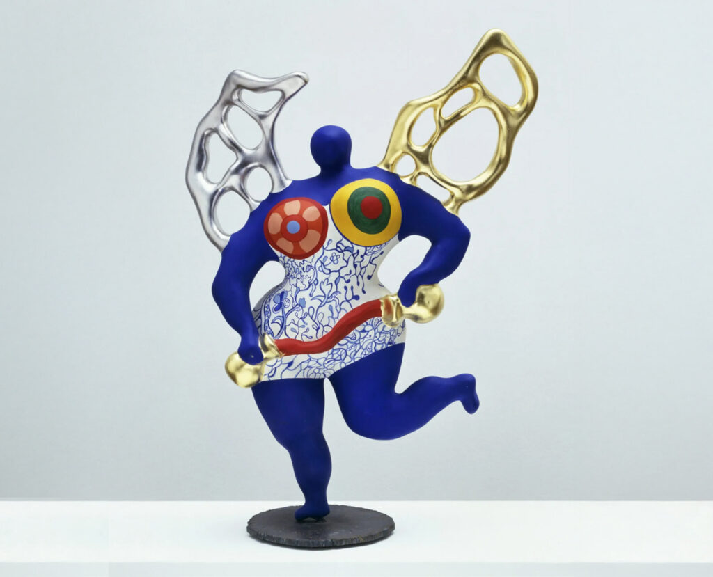Farbenfrohe abstrakte Skulptur einer blauen menschlichen Figur mit goldenen und silbernen flügelartigen Formen und lebhaften, gemusterten Brustmotiven.