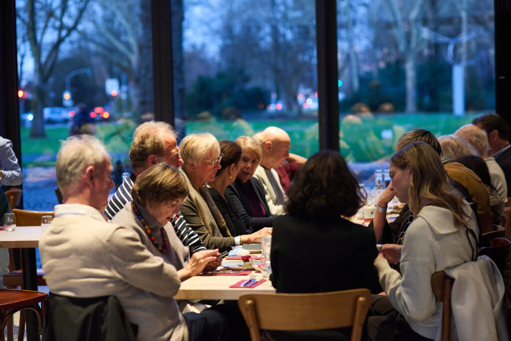 Eine Gruppe von Menschen unterschiedlichen Alters sitzt an einem langen Tisch in einem Restaurant und unterhält sich und unternimmt etwas.