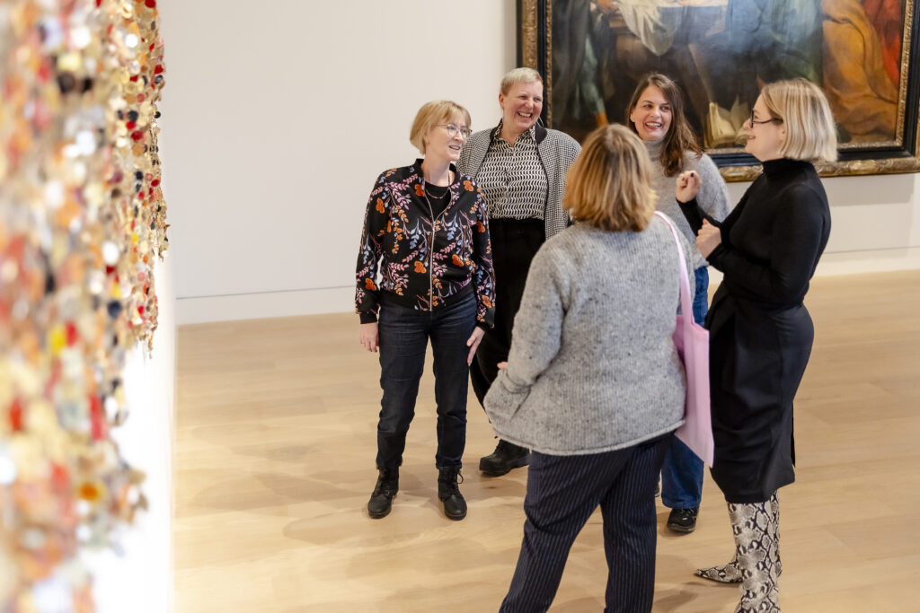 Fünf Frauen stehen lächelnd in einer Kunstgalerie und unterhalten sich in der Nähe einer bunten Wand und eines großen gerahmten Bildes.