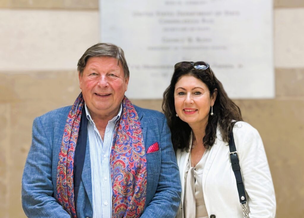 Ein lächelnder Mann in einer blauen Jacke mit einem bunten Schal steht neben einer Frau in einer weißen Jacke, die beide in einem Innenraum posieren.