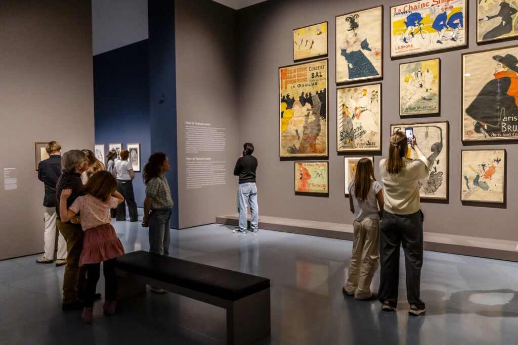In einer Galerie eines Kunstmuseums mit grauen Wänden und polierten Böden werden alte Plakate betrachtet und fotografiert.