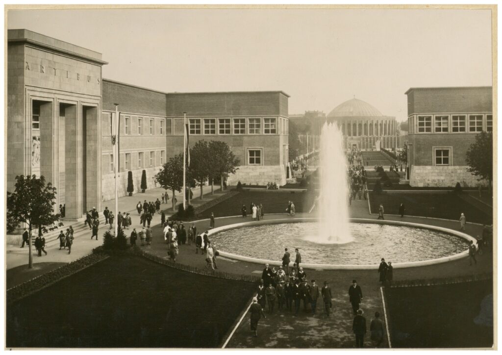 Schwarz-Weiß-Foto von Menschen, die um einen großen Springbrunnen vor großen Gebäuden in einer campusähnlichen Umgebung spazieren.