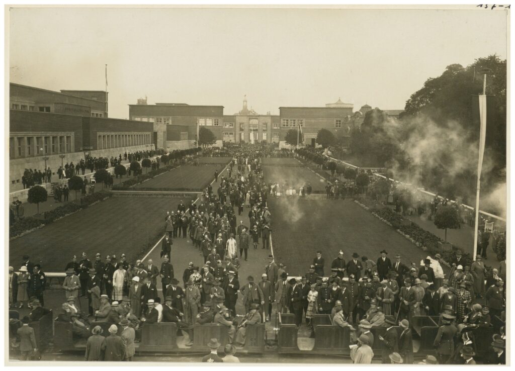 Eine große Menschenmenge versammelt sich in einem Gartenhof in der Nähe eines großen Gebäudes, wobei auf der rechten Seite des Bildes etwas Rauch zu sehen ist.