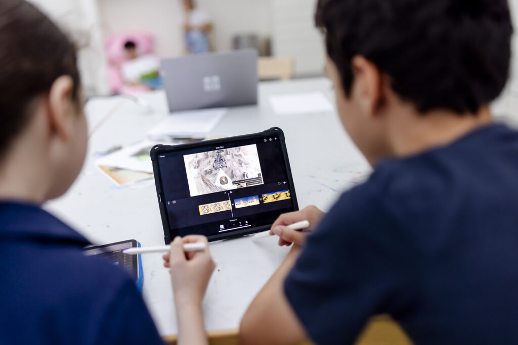 Zwei Kinder verwenden ein Tablet mit einem Stift und bearbeiten gemeinsam ein Video oder ein Bild an einem Tisch mit Laptops und Papieren in der Nähe.