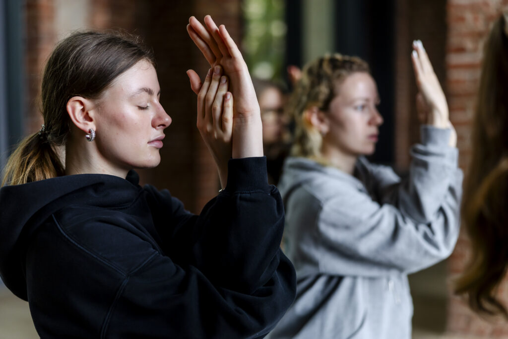 Zwei Frauen in Kapuzenpullis mit geschlossenen Augen heben ihre Hände an einem Ort im Innenbereich anmutig in einer meditativen oder tänzerischen Pose empor.