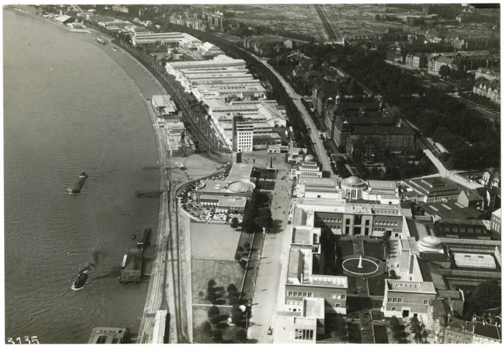 Luftaufnahme einer Stadt am Fluss mit Hafenanlagen, Booten, Industriegebäuden und grünen Parkanlagen entlang des Ufers.